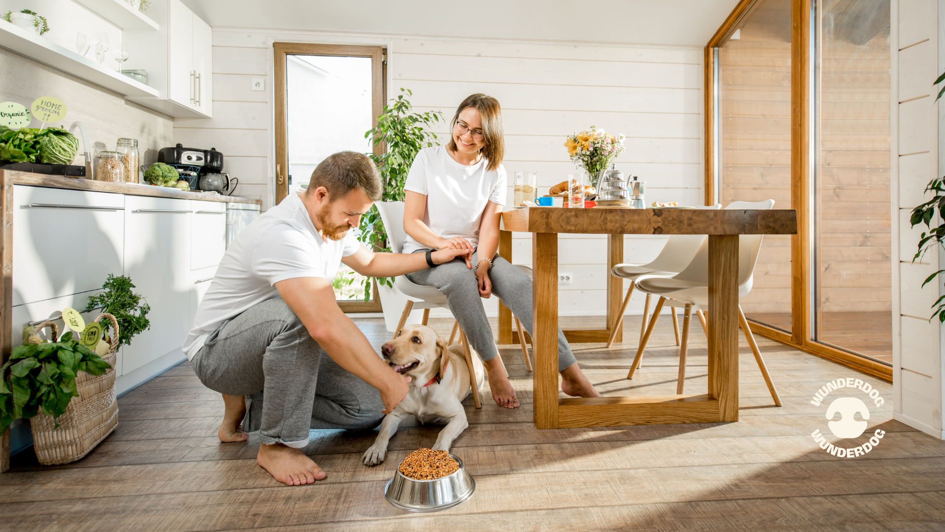 Happy couple in a bright kitchen bonding with their Labrador Retriever as it enjoys a bowl of Wunderdog fresh dog food, highlighting healthy feeding and allergy-friendly meals.