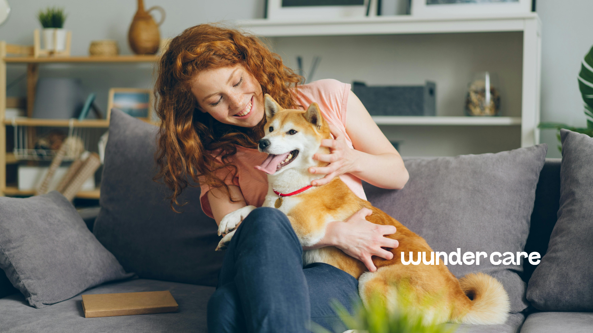 Photograph of a smiling woman on a grey sofa cuddling a happy Shiba Inu wearing a red collar. Bright indoor home setting, showing comfort and healthy skin under Wundercare logo.