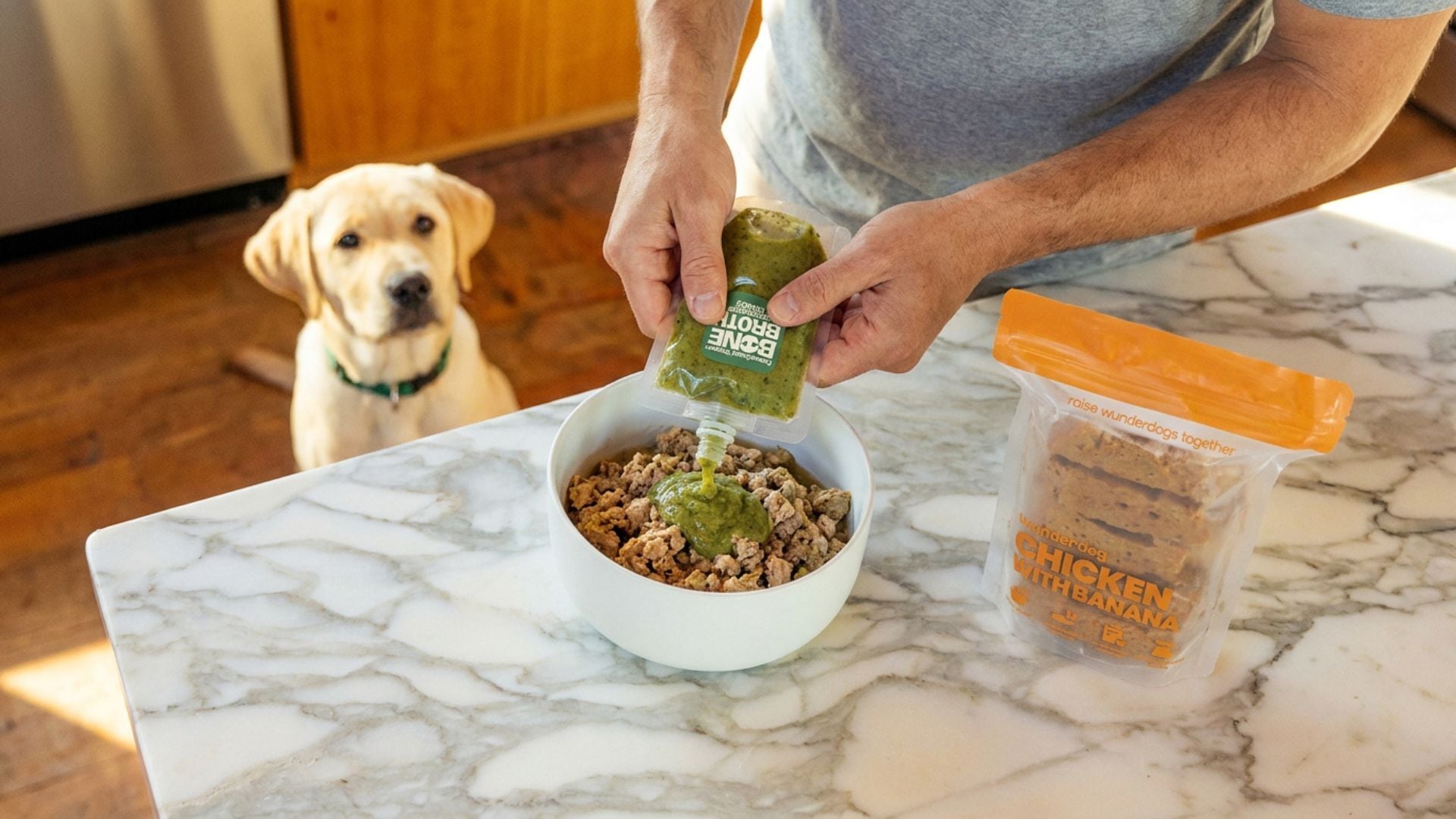 Alt text: Guardian pouring Supergreen Turkey bone broth topper over a bowl of Wunderdog Chicken with Banana meal on a marble kitchen counter, with a yellow Labrador puppy watching in the background